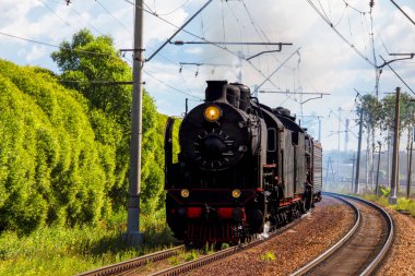 Retro buhar lokomotifi bacadan duman salıyor, demiryoluna çıkıyor, yeşil ağaçları geçiyor.