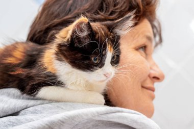 Küçük tüylü bir kedi yavrusu yaşlı bir kadının omzunda oturuyor.