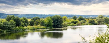 Nehir kenarındaki ağaçlarla ve bulutlu gökyüzüyle yaz manzarası, nehirdeki ağaçların yansıması.