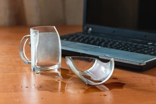Broken glass mug with spilled coffee or tea near laptop in the office