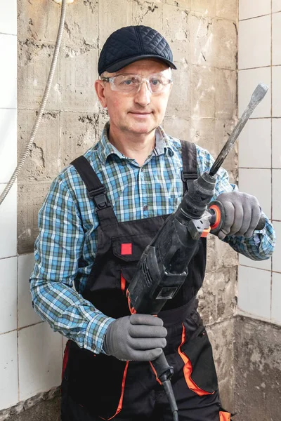 The craftsman in work clothes and protective glasses holds a perforator in his hands