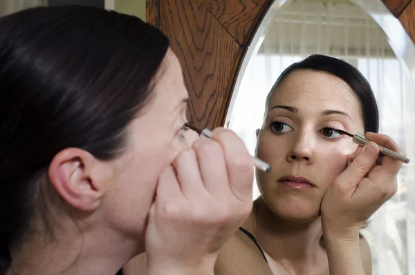 Jovem mulher olha no espelho e aplica eyeliner — Fotografia de Stock