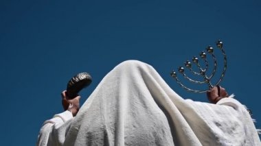 Yetişkin Yahudi bir adamın elinde iki Yahudi sembolü, bir menorah (R) ve bir shofar (ram boynuzu) (L) açık mavi gökyüzünün altında mesihin gelişini araştırıyor..