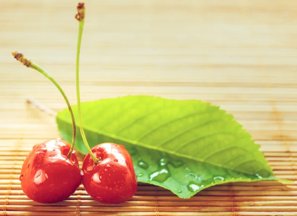 Two cherries placed near green leaf on bamboo tablecloth — Stock Photo, Image