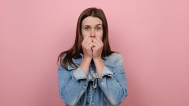Korkmuş, genç, esmer kadın 20 'li yaşlarda. Yüzünü parmaklarının arasından gizleyen kameraya bak. Kot ceket giyiyor, stüdyodaki pastel pembe arka plan duvarının üzerine izole edilmiş pozlar veriyor.