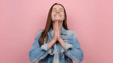 Yalvaran genç yirmili yaşlarda bir kadın dua ederken el ele tutuşup dilek dileniyor. Parmaklarını çapraz tutuyor, kot ceket giyiyor, stüdyoda pastel pembe arka planda izole edilmiş.