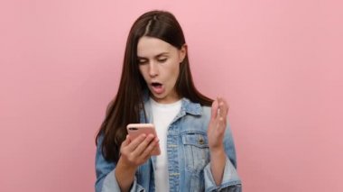 Cep telefonu kullanan mutlu genç esmer kadın evet dedi kazanan büyük bir haber yaptı kot ceket giymiş, pastel pembe arka plan duvarında izole edilmiş