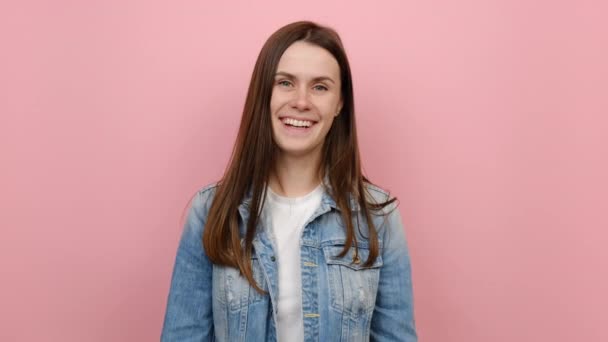 20岁的年轻的高加索女人穿着斜纹棉布夹克 高兴地看着相机 在演播室的粉刷粉红背景墙上摆出孤零零的笑容 人的生活方式概念 — 图库视频影像