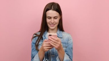 20 'li yaşlarda mutlu, mutlu bir kadın cep telefonu kullanıyor, cep telefonu kullanıyor, sohbet ediyor mavi kot ceket giymiş, pastel pembe arka plan stüdyosu üzerinde izole edilmiş poz veriyor, mesajlar yolluyor.