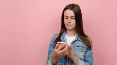 Mutlu, genç bir kadın cep telefonuyla fotoğraf çekiyor sosyal ağda sohbet ediyor, mavi kot ceket giyiyor, stüdyodaki pastel pembe arka plan duvarında izole edilmiş poz veriyor.
