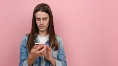 Genç milenyum kadınları cep telefonlarını tutarken el ile çalışma alanının fotokopisini çekiyorlar, kot ceket giyiyorlar, stüdyodaki pastel pembe arka plan üzerine izole edilmiş pozlar veriyorlar.