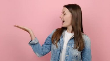 20 'li yaşlarda genç esmer bir kadın, kot ceket giyiyor. İşyeri fotokopi stüdyosunun reklam alanındaki yerlerini işaret ediyor. Kot ceket giyiyor, pastel pembe arka plan stüdyosunda izole edilmiş.