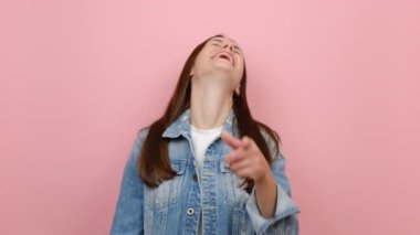 Heyecanlı, neşeli, esmer, 20 'li yaşlarında bir kız. Bak, gülümse, gülümse, komedi filmi işaret parmağı, kot ceket giy, pastel pembe arka plan stüdyosunda izole ol.