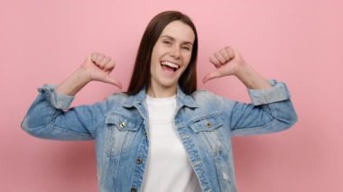 Kendinden emin, genç, beyaz, 20 'li yaşlarda, kendini işaret eden, başparmağını jest gibi gösteren, kot ceket giyen, stüdyodaki pastel pembe arka plan duvarının üzerine izole edilmiş bir poz veren bir kadın.