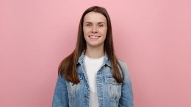 Gülümseyen, sevimli, arkadaş canlısı, 20 'li yaşlarda mutlu, kamera karşısında göz kırpan, kot ceket giyen, pastel pembe arka plan stüdyosunda yalnız poz veren bir kadının portresi. Duygu kavramı