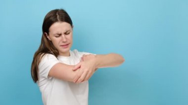 Mutsuz, üzgün, 20 'li yaşlarda beyaz bir kadının dirseğinde ağrı var. Travma üstüne yaralı elleri ısıtıyor. Arka plandaki mavi renkli duvar stüdyosunda fotokopi alanı ile izole edilmiş pozlar veriyor.