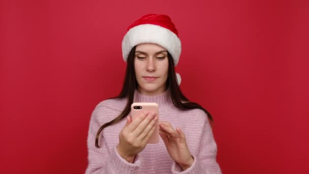 ポートレートのメリー若いサンタ白人女性ホールド使用携帯電話タイピング言うすごいはいちょうど大きな勝利のニュースを発見しました ピンクのセーターとクリスマスの帽子を身に着けています — ストック動画