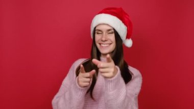 Heyecanlı, genç, 20 'li yaşlarda, pembe örgü süveter ve Noel şapkalı genç bir Noel Baba kadın kırmızı stüdyo arka planında izole edilmiş bir poz veren bir jest gibi başparmağını kaldırdığını gösteriyor. Mutlu Yıllar Kutlama Konsepti