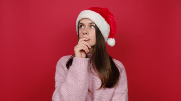 パズル陽気な若いサンタCaucasian女性20年代のルックス周り考えが出てくるアイデアを提起指 ピンクのニットセーターとクリスマスの帽子を身に着けています モデルは赤い色の背景スタジオ上にポーズ — ストック動画