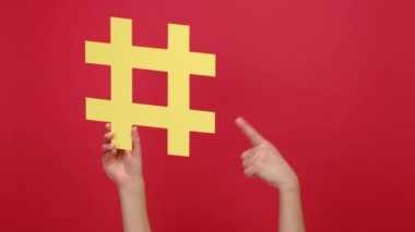 Kadın el, sarı hashtag 'i tutuyor ve parmağıyla işaret ediyor. Stüdyonun kırmızı arka plan duvarında izole edilmiş bir işaret var. Sosyal medya pazarlama sembolü, blog yazma, medya planlama kavramı
