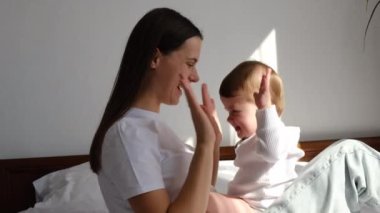 Sevecen, genç, beyaz bir annenin, tatlı, küçük kızıyla evde rahat bir yatakta otururken, hassas bir anın tadını çıkarması. Mutlu anne, gülen küçük bebekle vakit geçiriyor.