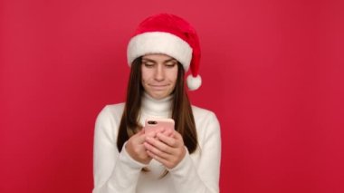Üzgün genç bir kadın cep telefonunda SMS yazıyor, beyaz örgü süveter ve Noel Baba şapkası giyiyor, kırmızı stüdyo arka planında izole edilmiş. Mutlu yeni yıl kutlamaları, mutlu tatiller.
