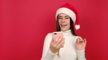 Mutlu genç bayan telefonu tut ve sanal Xmas konuşmasını kaydet. Noel şapkalı mutlu kadın akıllı telefon kullanarak selfie video konferansı yapıyor, kırmızı arka planda izole edilmiş. Sosyal uzaklık ve Yeni Yıl