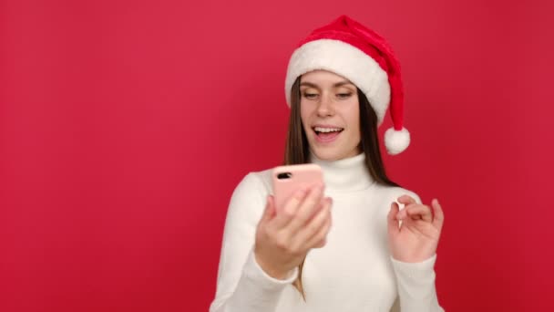 快乐的年轻女士拿着电话录下虚拟的圣诞讲话 戴圣诞礼帽的快乐女性使用智能手机进行自拍视频会议 用红色背景隔离 社会疏离与新年 — 图库视频影像