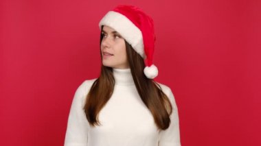 Gizli esmer kadın sessiz ol diyor dudaklarında şşşş jest, süveter ve Noel Baba şapkası giyiyor, kırmızı stüdyo arka planında izole edilmiş. Yeni yılı kutluyoruz. Mutlu tatiller.