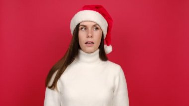 Beyaz kazaklı ve Noel Baba şapkalı kazanan genç kadın kulak misafiri olduğunu duymaya çalışıyor. Kırmızı stüdyo arka planında izole edilmiş. Mutlu yıllar tatil konsepti