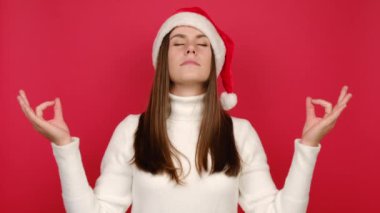 Sakin genç bir kadın yoga jestinde el ele tutuşur sakinleşmeye çalışırken meditasyon yapar, örgü örer ve Noel Baba şapkası giyer, kırmızı stüdyo arka plan duvarında izole olur. Mutlu yıllar, mutlu yıllar.