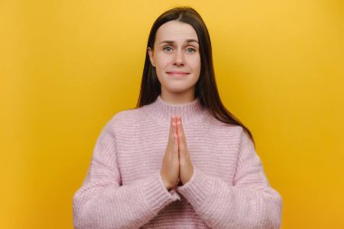 Yalvarılan portreyi kapat. 20 'li yaşlardaki üzgün, beyaz bir kadına dua jesti yapıp pembe örülü süveter, sarı stüdyo arka planında izole edilmiş bir şey için yalvarmasını söyle.