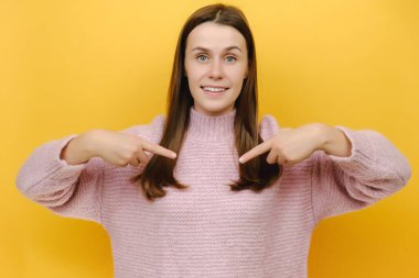 Gülüşü şaşkın, genç, şaşkın, esmer kadın işaret parmağı kameraya bak, rahat pembe örgü süveter giyiyor, stüdyonun sarı arka plan duvarında izole edilmiş.