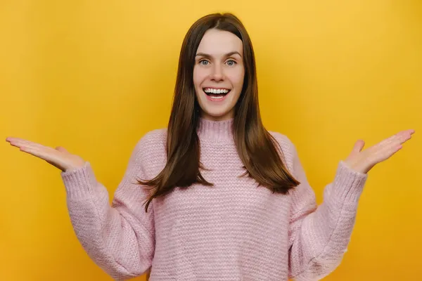 令人惊讶的是 令人惊讶的是 这位快乐的年轻高加索女人张开了双手 穿着粉色针织毛衣 在工作室中将自己隔离在黄色的背景墙之外 人的生活方式概念 — 图库照片
