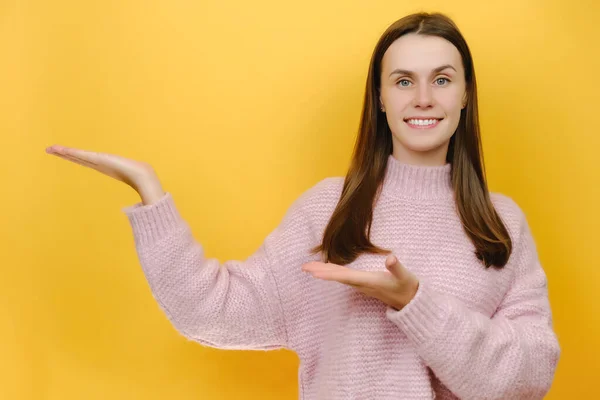 笑容可亲漂亮漂亮的千禧年女性穿着粉色针织毛衣 把食指放在工作区复制空间的模型上 在工作室的黄色背景墙上摆出隔离的姿势 — 图库照片