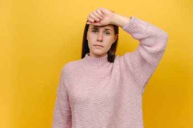 Yorgun, hasta, bitkin, genç, beyaz bir kadın elini alnına koydu kameraya baktı, baş ağrısı vardı, pembe örgü süveter giyiyordu, sarı arka plan stüdyosunun üzerinde izole bir şekilde poz veriyordu.