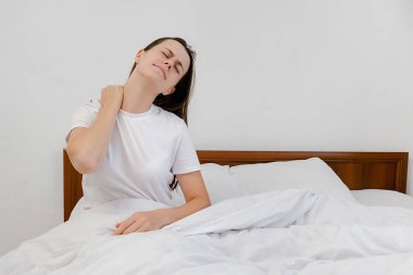 Fibromiyalji ve sırt ağrısı konsepti. Mutsuz genç bir kız rahatsız bir yatakta uyanıyor sırt ağrısı çekiyor gergin boyun kaslarına masaj yapıyor kötü bir yatakta uyuduktan sonra yanlış bir duruşla.