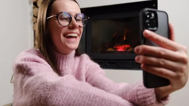 スマートフォンを使用して暖炉のある豪華なリビングルームで床に座っている眼鏡の笑顔の若い女性 話して録音ビデオやソーシャルネットワークでオンラインストリーミング 仮想イベントコンセプト — ストック動画