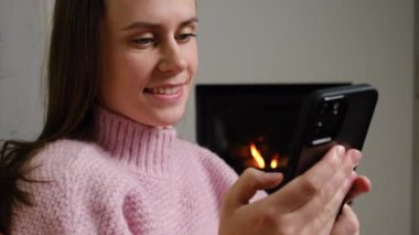 Güzel, güzel esmer kadının yakın portresi elinde akıllı bir telefon, şöminenin yanındaki kır evinde dinleniyor, e-ticaret mağazası sitelerinden alışveriş yapıyor, internet konseptinde tasasız hafta sonunun keyfini çıkarıyor.