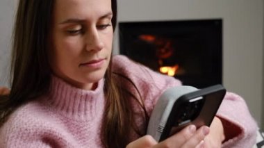 Güzel, genç ve güzel bir kadının portresi. Sıcak zeminde oturmuş şöminenin yanındaki yazlık evde dinleniyor. Akıllı telefon cihazı kullanıyor, internette sohbet ediyor, internetten alışveriş yapıyor.