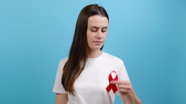概念世界エイズとHivの日 若いブルネットの女性は 青い色の背景スタジオに隔離されたカメラを見て 赤いリボンを保持しています 輸血と寄付 血友病と世界保健の概念 — ストック動画