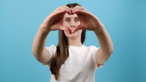 幸せな若い女性を閉じる指の心の形の笑顔でシンボルを介してカメラを見て 青の背景に孤立ポーズを作る クライアントは肯定的なフィードバック 健康保険 寄付の概念を与える — ストック動画
