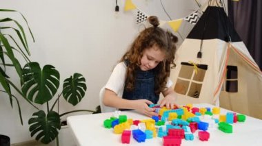 Kıvırcık kız, rahat oturma odasındaki beyaz masada plastik küplerle oynuyor. Sevimli küçük güzel çocuk oyun zamanı inşaatından zevk alıyor renkli tuğlalar kullanıyor. Hafta sonu eğlence etkinliği konsepti