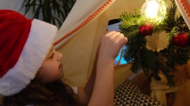 Kırmızı şapkalı sevimli kız çocuğu küçük bir Noel çelengi süslüyor çadırda oturuyor, kış tatili için evi süslüyor, kutlama için suni köknar takıyor, kapalı alanda kutlama atmosferi yaratıyor.