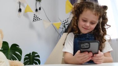 Çocuk kız akıllı telefonuyla masada oturup mobil uygulamalar kullanarak eğleniyor, evde oyunlar oynuyor. Cep telefonuyla öğrenen, video izleyen, mobil teknoloji konseptiyle eğlenen sevimli bir çocuk.