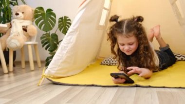Güzel kız çadırda akıllı telefonuyla yatıyor, mesajla kurye gönderiyor, internette oyun oynuyor. Eğlence için modern teknoloji. Siber ortamda çocukların güvenliği için ebeveyn denetim yazılımı