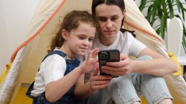 Mutlu genç bir anne, küçük şirin kızına akıllı telefon kullanarak öğrenme öğretiyor. Telefona bakıyor, mobil oyunlar oynuyor, teknoloji eğitimi için uygulamalar kullanıyor. Evdeki rahat çadırda oturuyor.