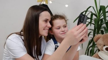 Modern teknolojiye bağımlı mutlu anne ve tatlı kız cep telefonu kullanarak eğleniyor. Sevimli, genç bir dadının sevimli küçük bir kızla mobil oyunlar oynaması ve evinde oturması.