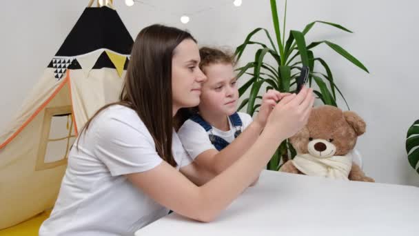 若いブルネットの母親は小さなかわいい子供の娘を教えているスマートフォンを使って学ぶスマートフォンを使って携帯ゲームを楽しんでいるハイテク教育エンターテイメントのためのアプリを使用してテーブルの中にリビングルームに座っている — ストック動画