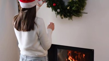 Noel Baba şapkalı, güzel, genç, beyaz kadın Noel çelengini yeni yıl süslemeleriyle süslemiş, evdeki şöminenin yanında duruyor. Xmas kış tatili konsepti